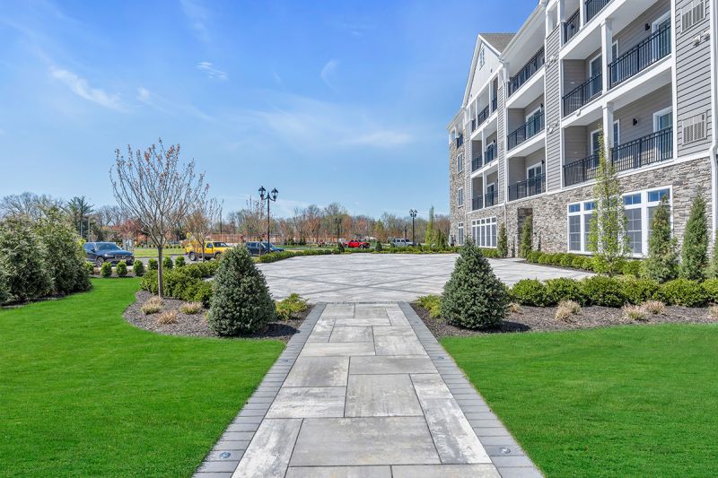 Exterior photo of an outdoor patio area at the Enclave at Woodhaven showing a large paver stone area surrounded by lush landscaping and easy access to the apartment building.