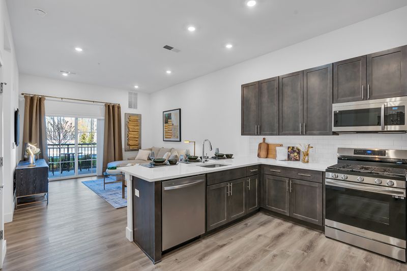 Interior photo of an apartment at the Enclave at Woodhaven showing the kitchen with luxury vinyl wood grain flooring, dark brown cabinets with white granite countertops and stainless steel appliances.\, the living room with balcony access can be seen in the photo.