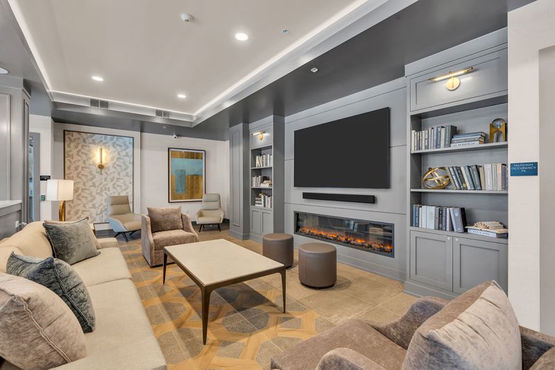 Interior photo of the resident lounge at the Enclave at Woodhaven showing several comfortable seating areas in front of a fireplace with large screen tv above. Built in book shelves with books flank both sides of the fireplace.