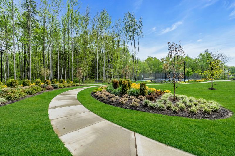 Exterior photo of the park at Enclave at Woodhaven showing a curved concrete walking path that runs through the community. On both sides of the path show neatly landscaped garden beds filled with a mix of mulches. There are multiple black lampposts and benches scattered in the field. A fenced-in tennis court is visible in the background.