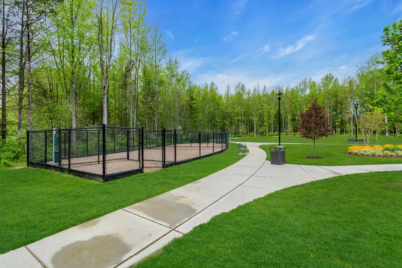 Exterior photo of the park at Enclave at Woodhaven showing a curved concrete walking path that runs through the community. To the left of the path is a large fenced-in area. There are multiple black lampposts and benches scattered in the field.