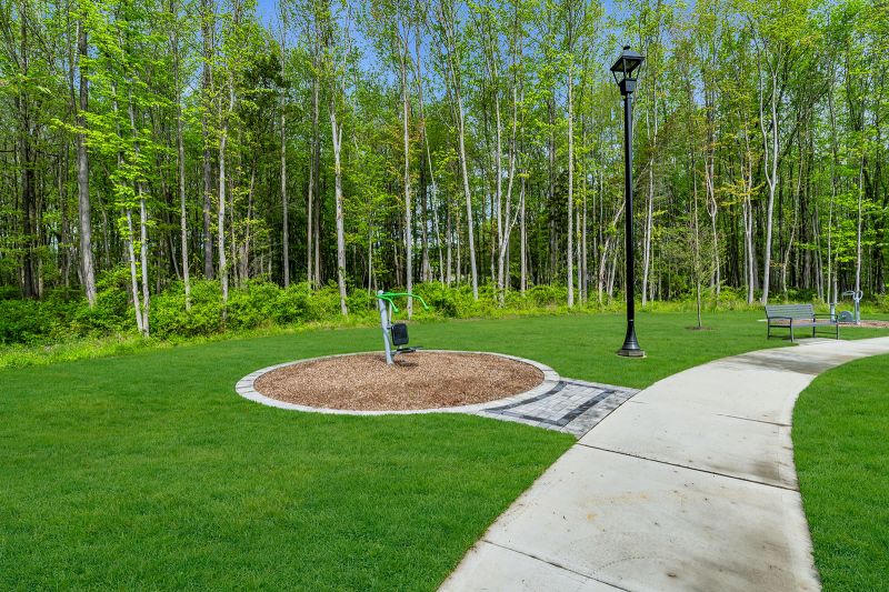 Exterior photo of the park at Enclave at Woodhaven showing a curved concrete walking path that runs through the community. To the left of the path, a piece of outdoor fitness equipment is installed on a circular mulch surface surrounded by a border of stone pavers. A tall black lamppost stands nearby, and a metal bench is placed along the path on the right. In the background, another exercise station and small tree are visible