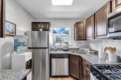 Interior photo of the kitchen at Carteret Garden showing cherry cabinets, stainless steel appliances, granite countertops, and luxury LVT woodgrain flooring. A small window allows natural light and views of the rest of the community.