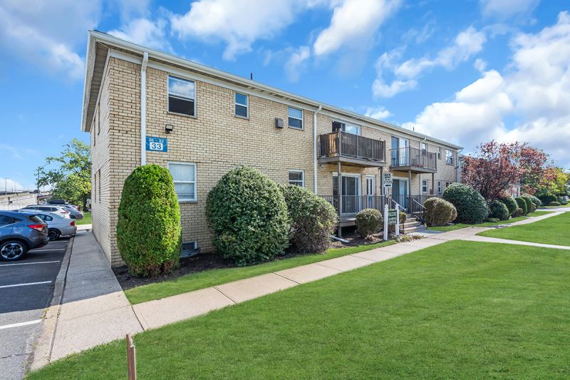 Exterior photo of the leasing office at Carteret Garden showing the exterior finishes including white brick with white trims, windows, and balconies with wood railings. A walkway with steps leading to the leasing office. An open parking lot with directional signage can be seen on the left. Green grass and bushes highlight the surrounding landscaping.