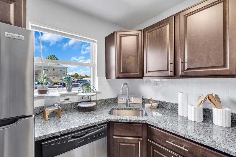 Interior photo of the kitchen at Carteret Garden showing cherry cabinets, stainless steel appliances, granite countertops, and luxury LVT woodgrain flooring. A large window allows natural light and views of the rest of the community.