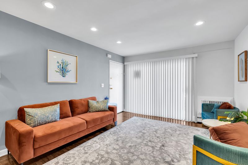 Interior photo of the living room at Carteret Garden showing the front door leading to a large couch, two side chairs with a coffee table, and luxury hardwood flooring. Sliding doors with vertical window blinds can be seen next to the front door.
