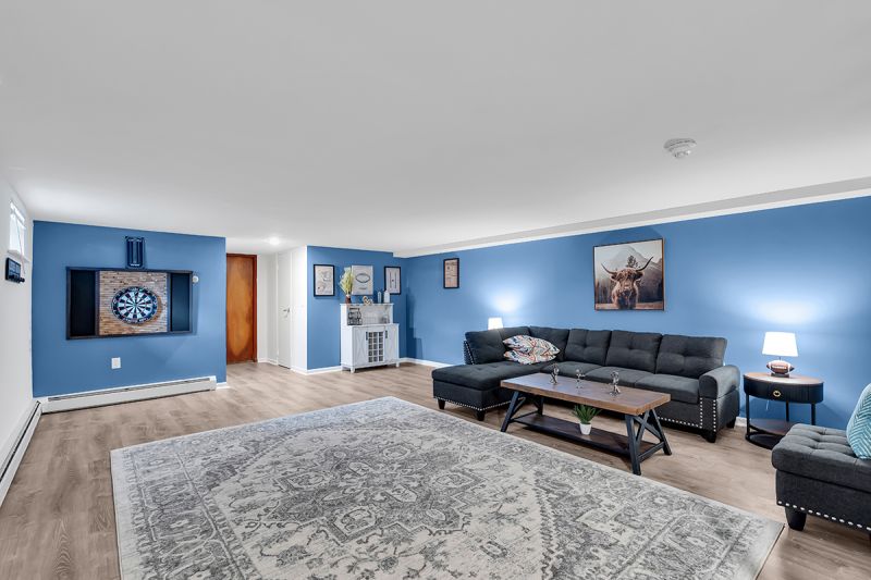 Interior photo of the recreation room at Carteret Garden showing luxury hardwood flooring, a large couch with a coffee table in front, two side tables with lamps, and a sofa chair for extra seating. A dartboard game can be seen on the wall.