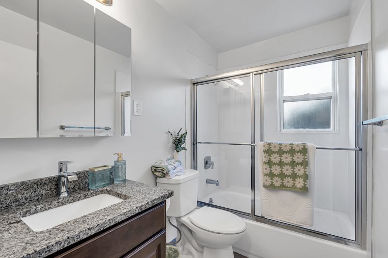 Interior photo of the bathroom at Carteret Gardens showing a tub with glass sliding doors, a toilet, and a large vanity with granite counter, cherry cabinets, and a medicine cabinet with attached mirror.