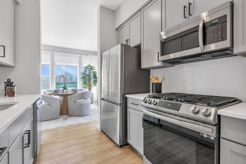Interior photo of kitchen at Woodland Square showing modern stainless-steel appliances, LVT flooring, grey cabinets, and white Quartz countertops with white subway-tiled backsplash and an open view of the formal dining room.