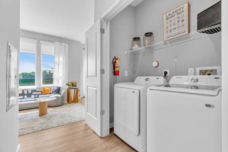 Interior photo of a large closet in Woodland Square showing side-by-side full-size washer and dryer with shelving above and lighting inside. There are double doors to conceal the machines when not in use.
