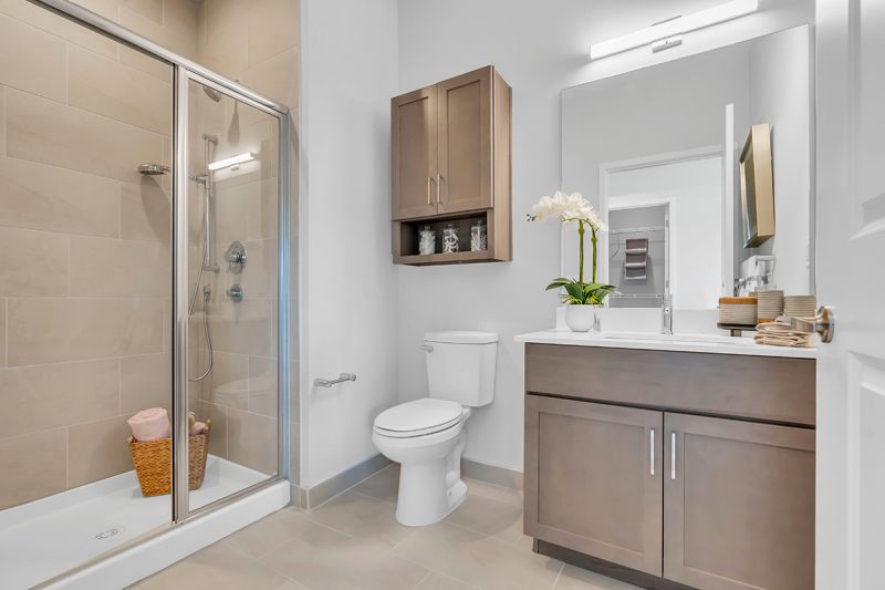 Interior photo of the primary bathroom at Woodland Square showing ceramic tile flooring, white quartz countertops, and light-colored vanity, large mirror. To the left of the vanity is a stand-up shower with a sliding door, and a toilet with a medicine cabinet located above.