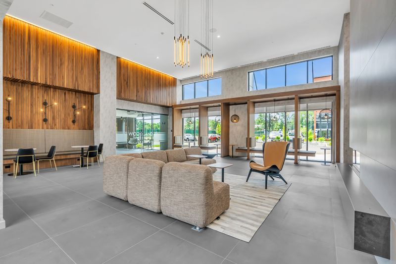 Interior photo of the community lounge at Woodland Square featuring a large sectional couch with a chair and two end tables in the center of the room. To the right are two-person seating cubicles with overhead light fixtures and large floor-to-ceiling windows. In the background, there is additional cafe seating with tables, and entry doors to the leasing office are located on the right.