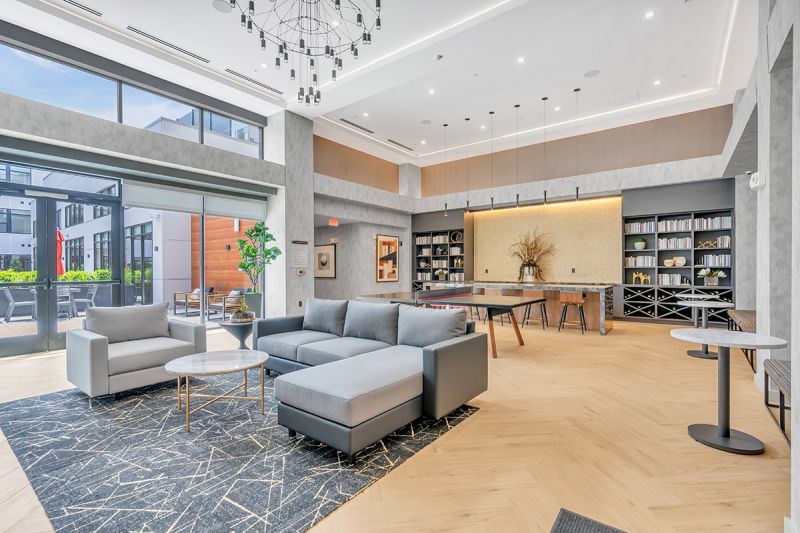 Interior photo of the community lounge at Woodland Square featuring a large sectional couch and a coffee table. Behind the couch is a ping pong table surrounded by a large marble counter with bar-height seating and mounted shelves with books and decorative pieces.