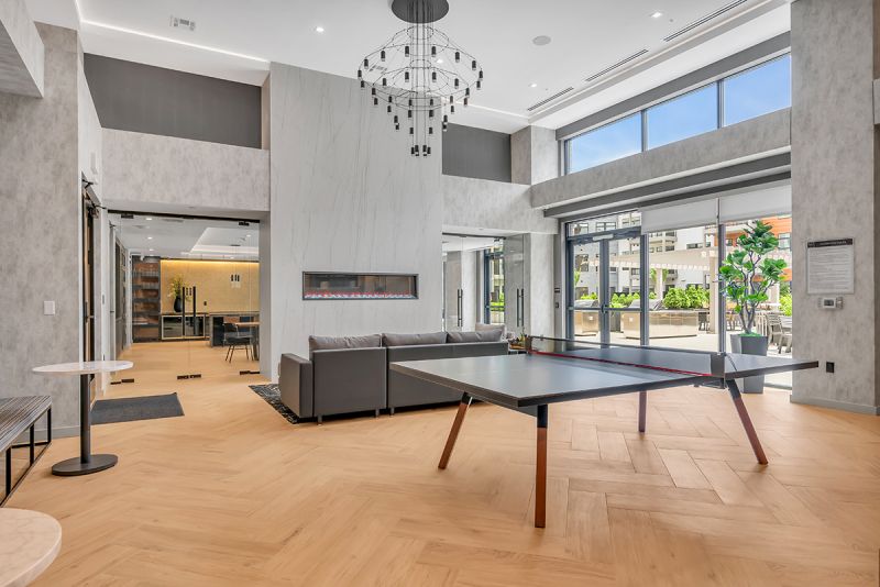 Interior photo of the community lounge at Woodland Square featuring a ping pong table in the center, and a large sectional couch that faces an electric fireplace. Along both sides of the electric fireplaces are double glass doors leading to the community room. The community’s exterior grill area is shown in the background.