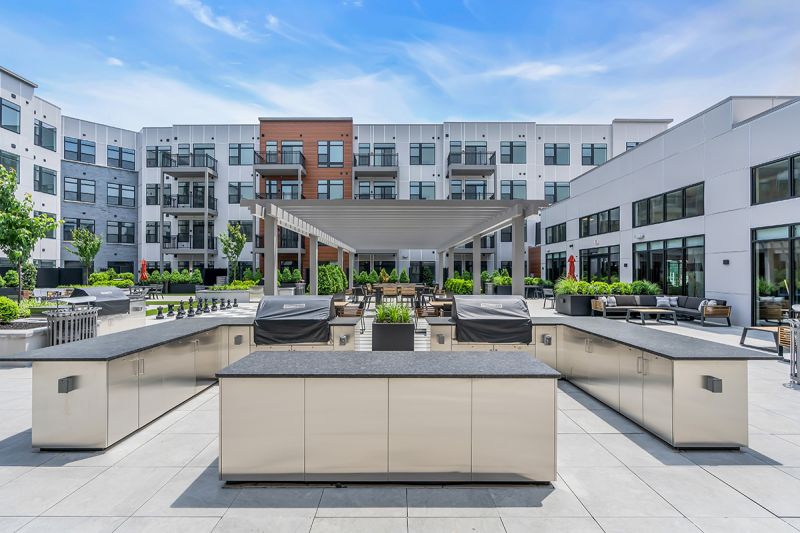 Exterior photo of the community grill at Woodland Square featuring two grills surrounded by a large marble counter with stainless steel cabinets overlooking additional features of the community’s exterior amenities.