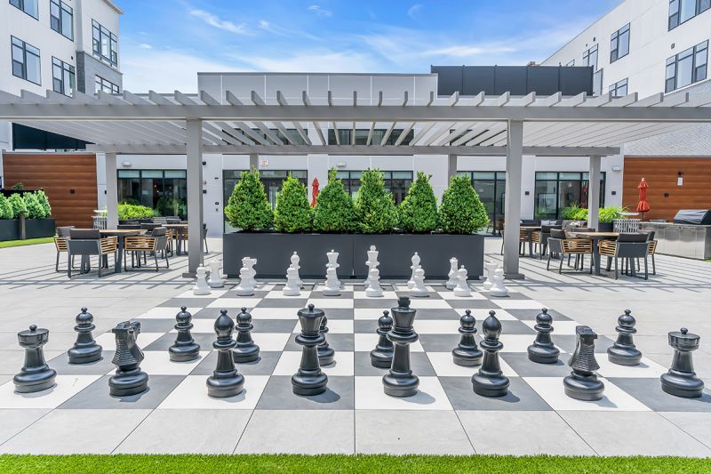Exterior photo of the community grill at Woodland Square featuring a giant chess game overlooking the seating area and grill surrounded by several bushes and trees.