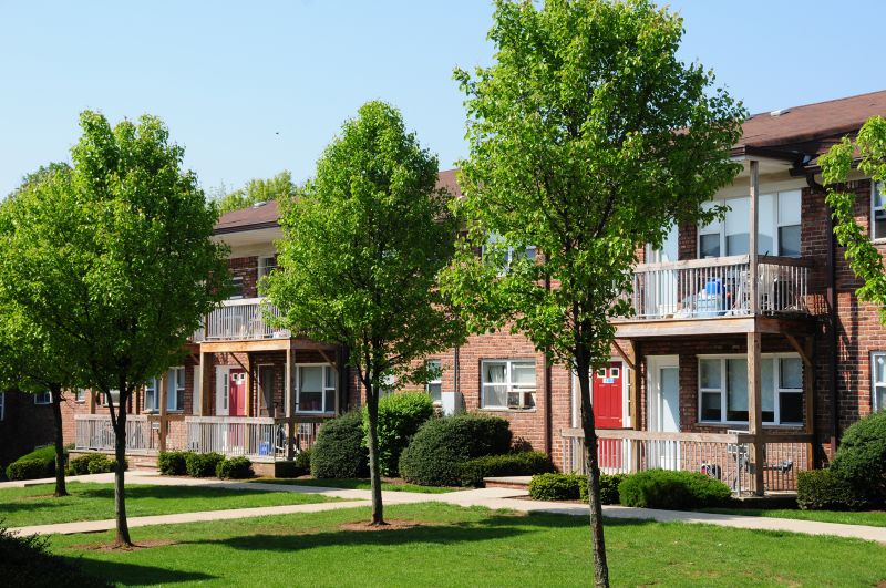Exterior photo of Hilltop Manor showing a brick apartment building with balconies. A lush green lawn with mature bushes and trees landscape the area.