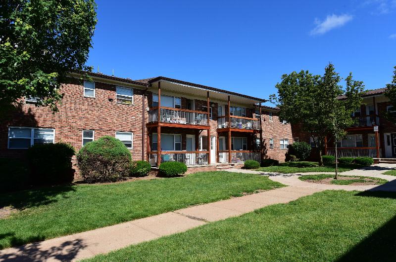 Exterior photo of Hilltop Manor showing a brick apartment building with balconies. A lush green lawn with mature bushes and trees landscape the area.