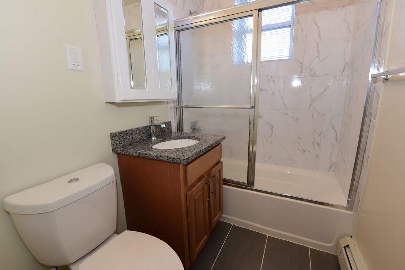 Photo showing a freshly painted renovated bathroom showing new ceramic tile flooring, new tub with shower doors, new toilet, sink, vanity and medicine cabinet.