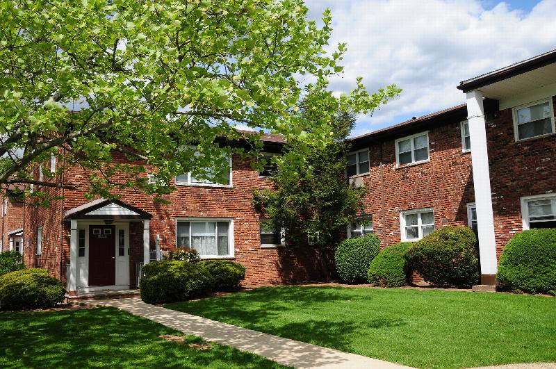 Hilltop Estates exterior photo showing a red brick apartment building with white trim. A lush green lawn with mature bushes and trees landscape the area. A sidewalk leads to the entranceway.