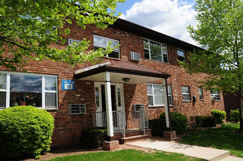 Hilltop Estates exterior photo showing a red brick apartment building with white trim. A lush green lawn with mature bushes and trees landscape the area.