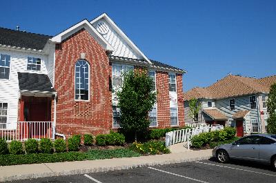 Rivendell Heights Exterior photo showing a brick and vinyl exterior. There is a pathway that leads to apartment entrances. Several bushes and trees highlight the landscape.