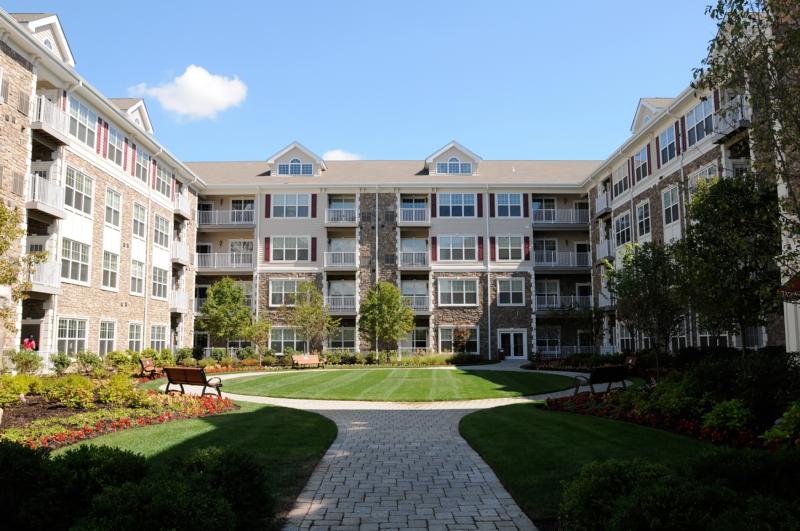 Helix at Xchange exterior photo shows lush landscaped court yard. Court yard is surrounded by a four story apartment building with stone and beige colored siding, red shutters and white trim. Center of court yard has circular concrete paved walkway lined with garden style shrubs, trees and park benches for seating. 