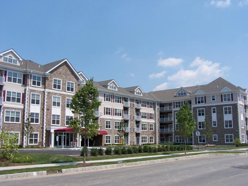 Helix at Xchange exterior photo shows the front lobby of a four story apartment building with stone and beige colored siding, red shutters and white trim. Exterior lobby is surrounded by green grass, shrubs and tree lined streets. 