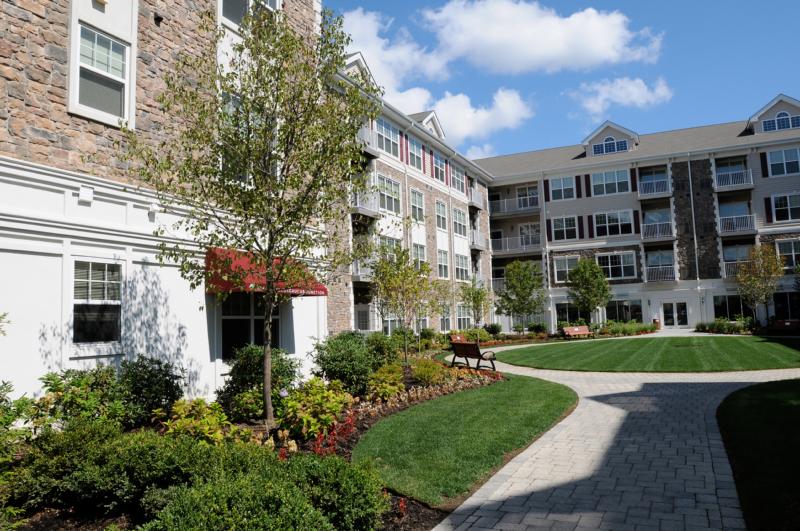 Helix at Xchange exterior photo shows a four story apartment building with stone and beige colored siding, red shutters and white trim. Landscaped court yard can be seen with garden style shrubs, trees, concrete paved walkway and park benches for seating.