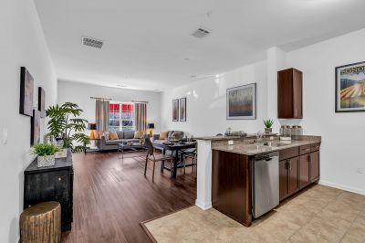 Interior photo of a model at Helix at Xchange showcasing the open floor concept of the kitchen and living room. The space features luxury hardwood flooring. The kitchen includes a bar-height counter, modern cabinetry, and a four-person dinette with a table and chairs. In the living area, two sofas are arranged around a central coffee table. The space features elegant decor and artwork including a large window to allow in natural light.