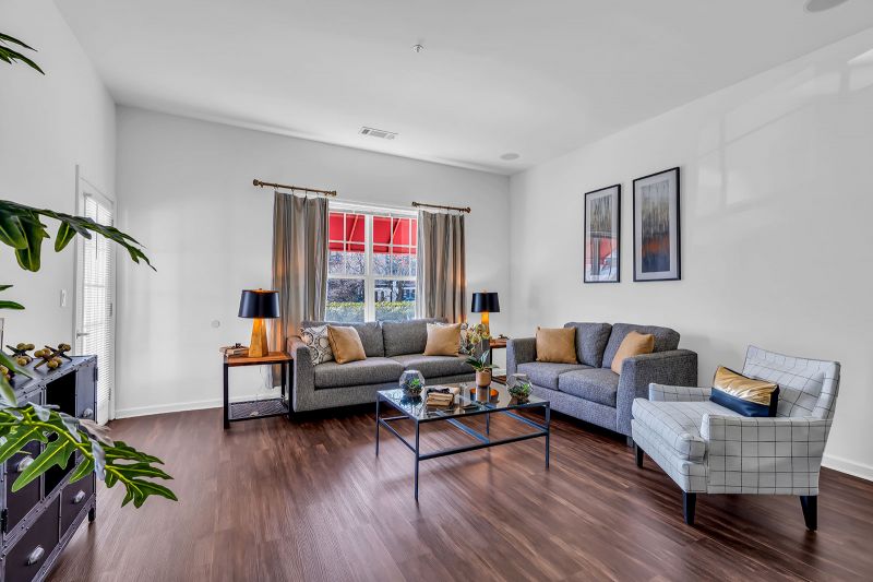 Interior photo of a model unit at Helix at Xchange showcasing the living room, which features luxury hardwood flooring. The space is furnished with two couches, a coffee table, an accent chair, and two side tables topped with lamps. Decorative pieces and artwork enhance the room's aesthetic, while a large window invites abundant natural light.