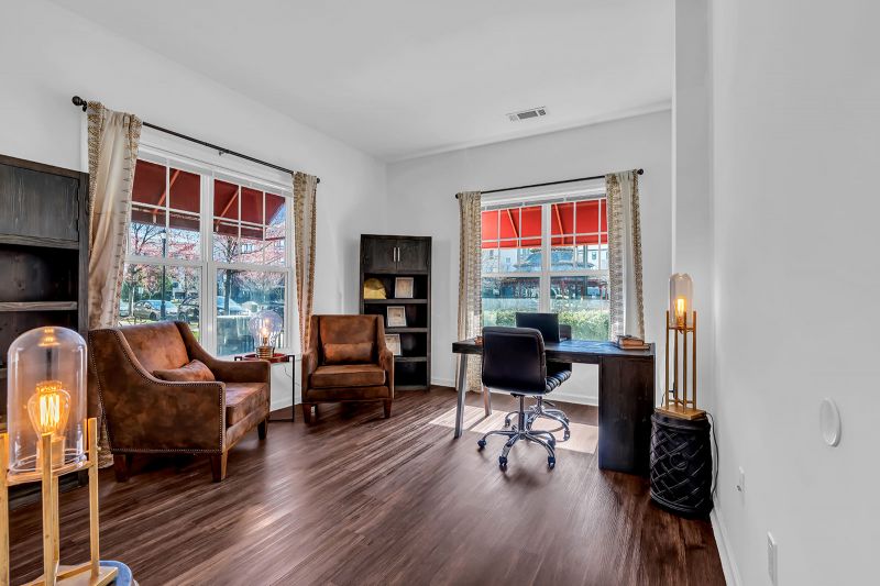 Interior photo of a model unit at Helix at Xchange showcasing a stylish home office with luxury hardwood flooring. The room features a desk with a rolling chair, two accent chairs for additional seating, and two large windows that fill the space with natural light.