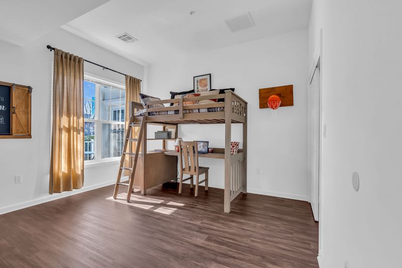 Interior photo of a model unit at Helix at Xchange showcasing an additional bedroom with luxury hardwood flooring. The room features a loft bed with an attached desk and chair, a closet with sliding doors on the right, and a large window that fills the space with natural light.