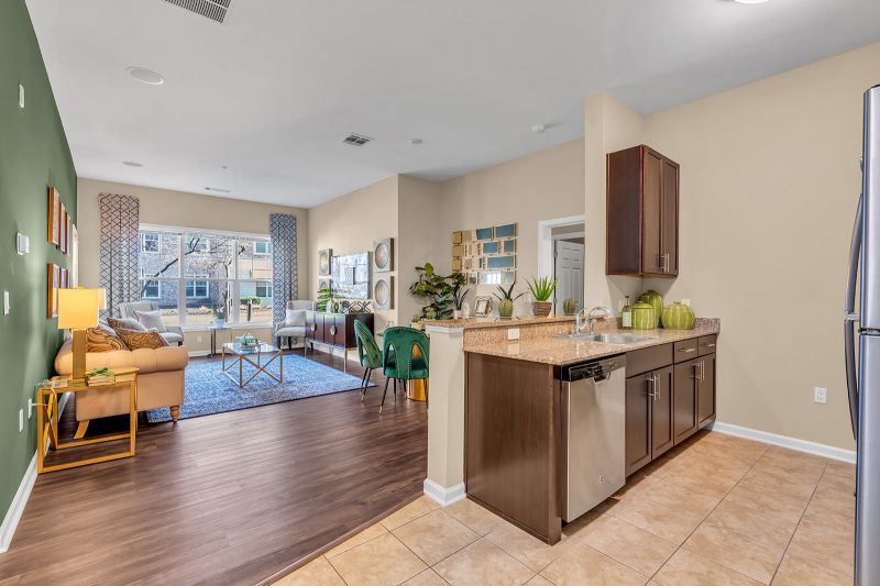 Interior photo of a model at Helix at Xchange showcasing the open floor concept of the kitchen and living room. The space is finished with luxurious hardwood flooring throughout. The kitchen features a bar-height counter, modern cabinetry, and a four-person dinette with a table and chairs. In the living area, a large couch and accent chairs are arranged around a central coffee table. Elegant decor and artwork complement the space, while a large window allows abundant natural light to fill the room.