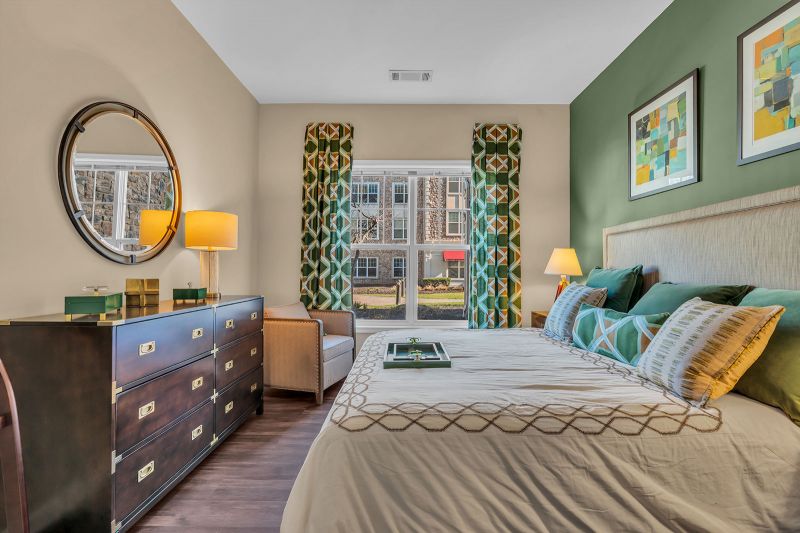 Interior photo of a model at Helix at Xchange featuring a bedroom with luxurious hardwood flooring. The room includes a bed with side tables on each side, an accent chair, and a dresser with a mirror above it. A large window lets in plenty of natural light, brightening the space.
