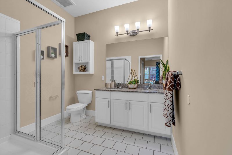 Interior photo of a model at Helix at Xchange showcasing a bathroom featuring sleek white porcelain tiles. The space includes new cabinetry, a double-sink vanity with a large mirror, a toilet with a cabinet above, and a stand-up shower. A unique light fixture above the mirror adds a stylish accent.