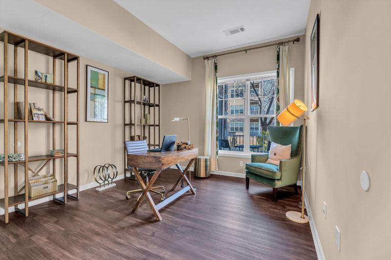Interior photo of a model unit at Helix at Xchange showcasing a stylish home office with luxury hardwood flooring. The room features a desk with a rolling chair, an accent chair for additional seating, and a large window that fills the space with natural light. Elegant decorative pieces and artwork add a refined touch to the room.