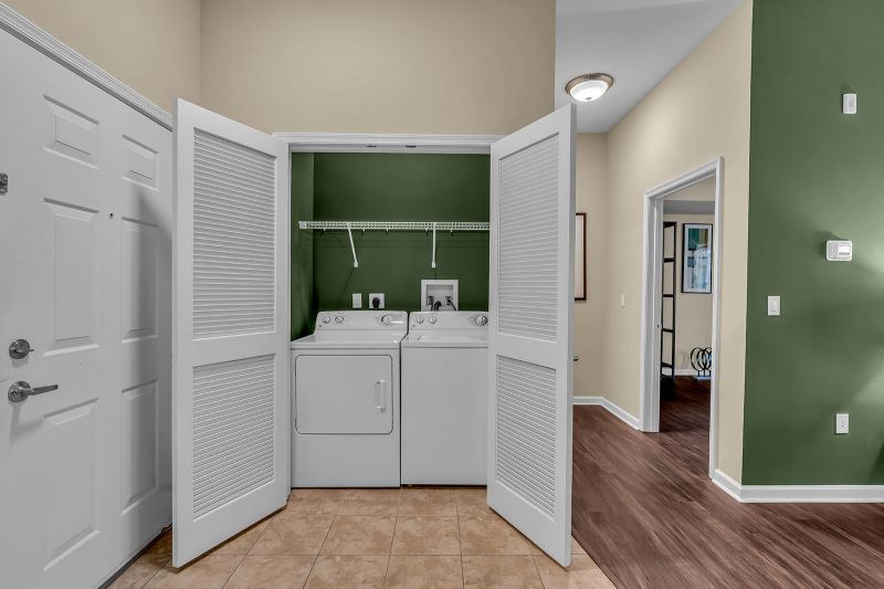 Interior photo of a model unit at Helix at Xchange showing the laundry area with a full-size washer and dryer inside a closet. The front entrance door and other areas of the unit are also visible in the frame.