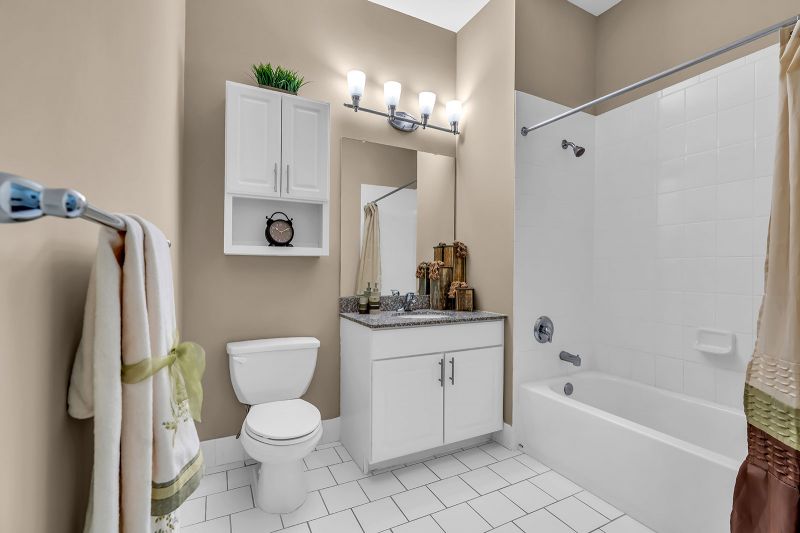 Interior photo of a model unit at Helix at Xchange showcasing a bathroom with modern white porcelain tiles. The space includes new cabinetry, a single-sink vanity with a mirror, a toilet with a cabinet above, and a shower-tub combo. A unique light fixture above the mirror enhances the room’s style.