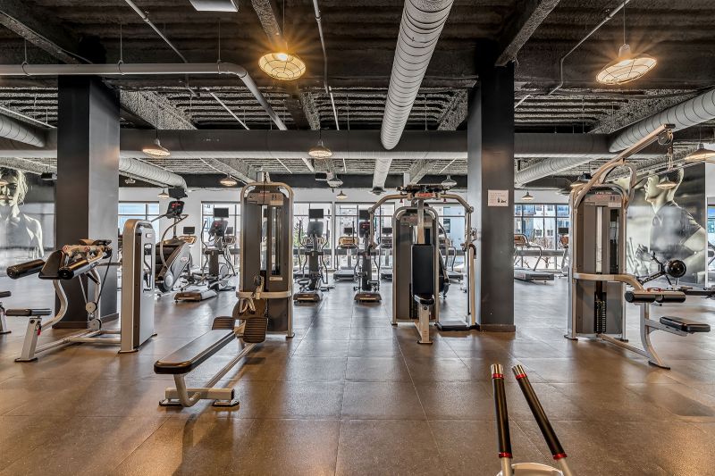 Interior photo of the gym at Xchange at Secaucus showing another angle of the variety of benches, squat racks, and lifting platforms to accommodate different workouts. Rubber flooring provides durability and safety, while large mirrors help users monitor their form. The space is brightly lit and neatly organized with storage racks.