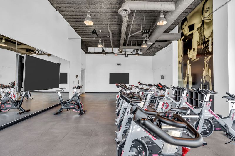 Interior photo of the spin room at Xchange at Secaucus featuring multiple stationary bikes arranged in rows, all facing large mirrors and a wall-mounted screens for virtual classes or guided workouts.