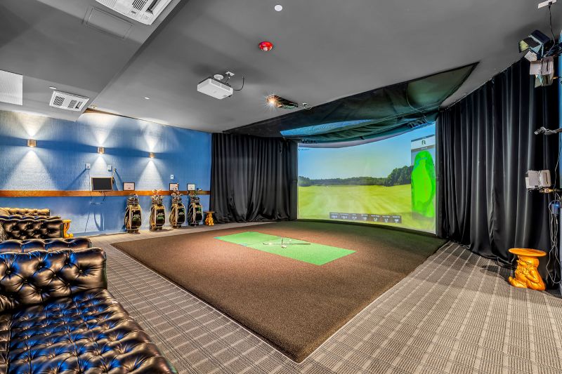 Interior photo of the golf stimulator room at Xchange at Secaucus showing a large wall-mounted projection screen displaying a variety of virtual courses, paired with realistic turf flooring, two golf clubs, and a golf ball. The room is equipped with comfortable seating, ample lighting, and club storage, creating a relaxing and functional space for both practice and entertainment.