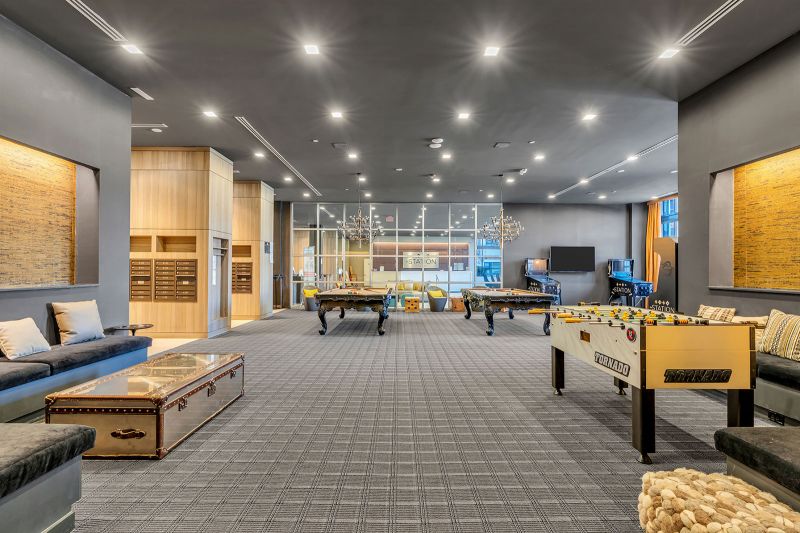 An interior photo of the game room at Xchange at Secaucus showing two pool tables, two arcade game machines, and a foosball table. The space includes multiple seating areas with couches, chairs, and tables, creating a comfortable setting for relaxation and socializing. A large, mounted TV adds to the entertainment experience, while mailbox cubbies for residents are visible on the right. Elegant light fixtures and a glass wall overlooking the lobby further enhance the room’s upscale ambiance.