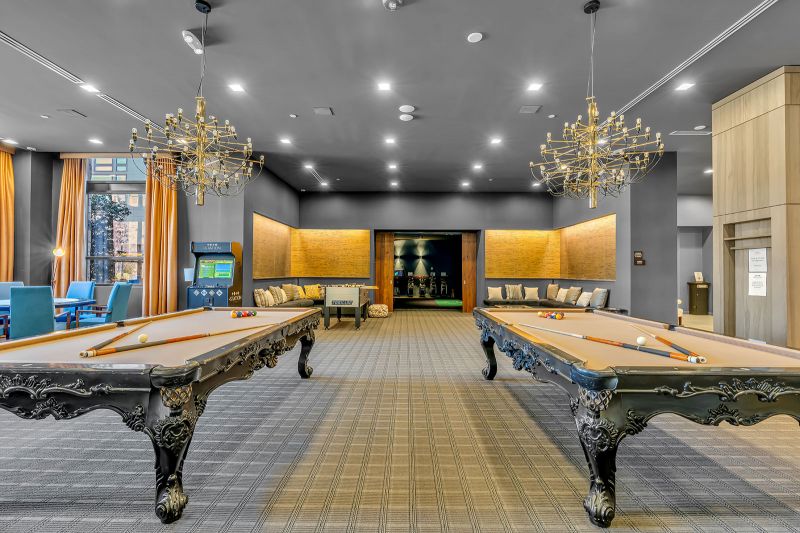 An interior photo of the game room at Xchange at Secaucus showing another angle of the two pool tables and foosball table. The space includes multiple seating areas with couches, chairs, and tables, creating a comfortable setting for relaxation and socializing. The entrance to the golf stimulator room can been seen in the background.
