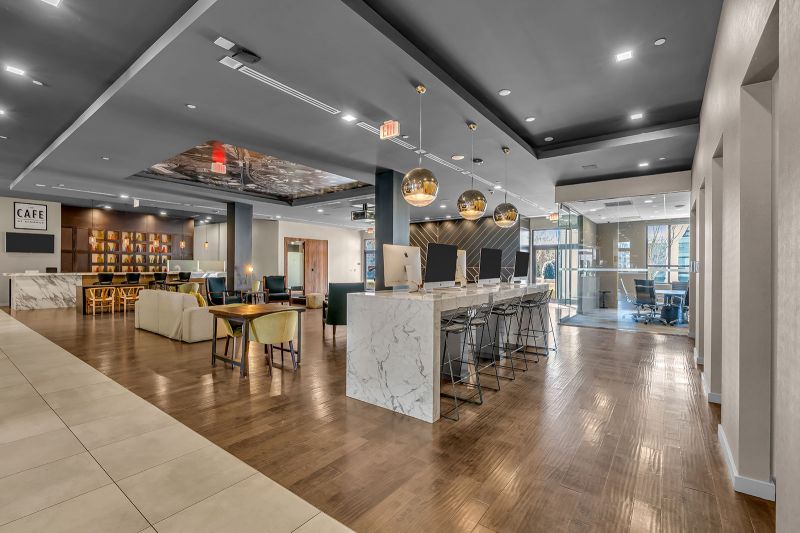 Interior photo of the lounge at Xchange at Secaucus showing luxury furnished seating areas, with stylish chairs and tables arranged in the center of the room. To the right, stationary computers with stools offer a convenient setup for remote work, while a café table with additional seating is positioned to the left. Multiple elegant light fixtures enhance the ambiance, creating a sophisticated and versatile space for both productivity and relaxation.