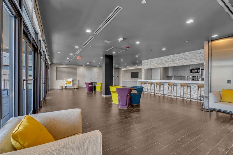 Interior photo of the event space at Xchange at Secaucus highlights the luxurious hardwood flooring and thoughtfully designed layout, perfect for hosting events. The space offers multiple seating areas with couches, chairs, and tables, creating a comfortable and inviting atmosphere. To the left, a fully equipped kitchen with bar-height counters and stools adds convenience and style. A large, mounted TV serves as the perfect addition for entertaining groups. Large windows overlooking the property flood the room with natural light.