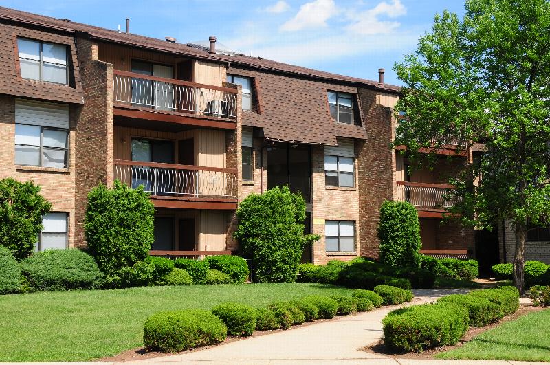 Blueberry Village exterior photo shows several entrances to a brick apartment building with balconies. Several trees and flowering bushes surround the area.
