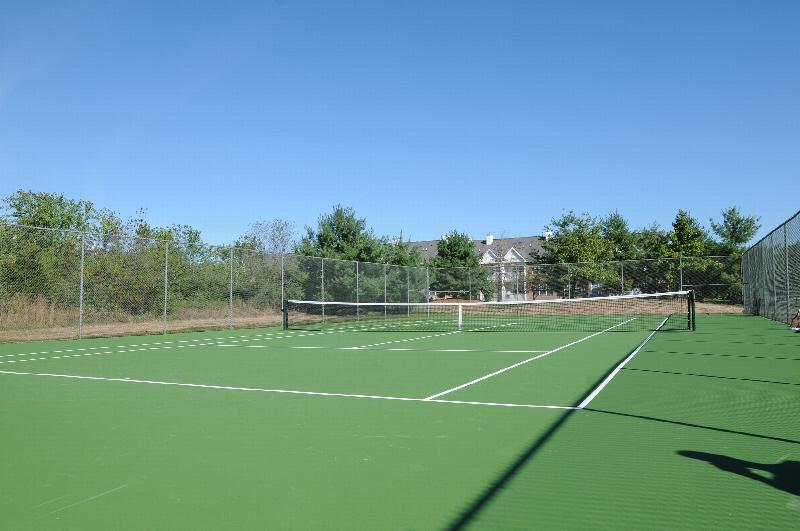 New Center Greens Tennis Court photo showing the tennis courts surrounded by a fence. The apartment building can be seen in the background. Several bushes and trees highlight the landscape.