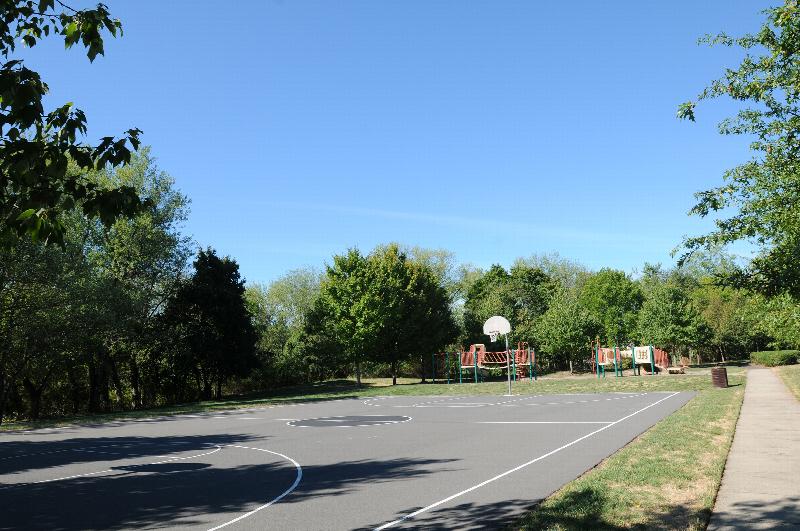 New Center Greens Basketball Court photo showing a full size basketball court. The playground can be seen in the background. Several bushes and trees highlight the landscape.