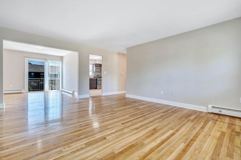 Interior photo of a refurbished apartment at Beekman Gardens in Hillsborough, NJ showing fresh paint, newly refinished floors leading from the living room to the dining room. Sliding glass doors to a balcony and an entrance to the kitchen can be seen.